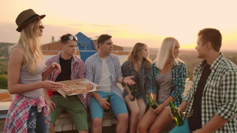 The-company-of-six-young-people-communicate-with-each-other-on-the-roof-with-pizza-and-beer.-Girls-look-around-at-summer-sunset